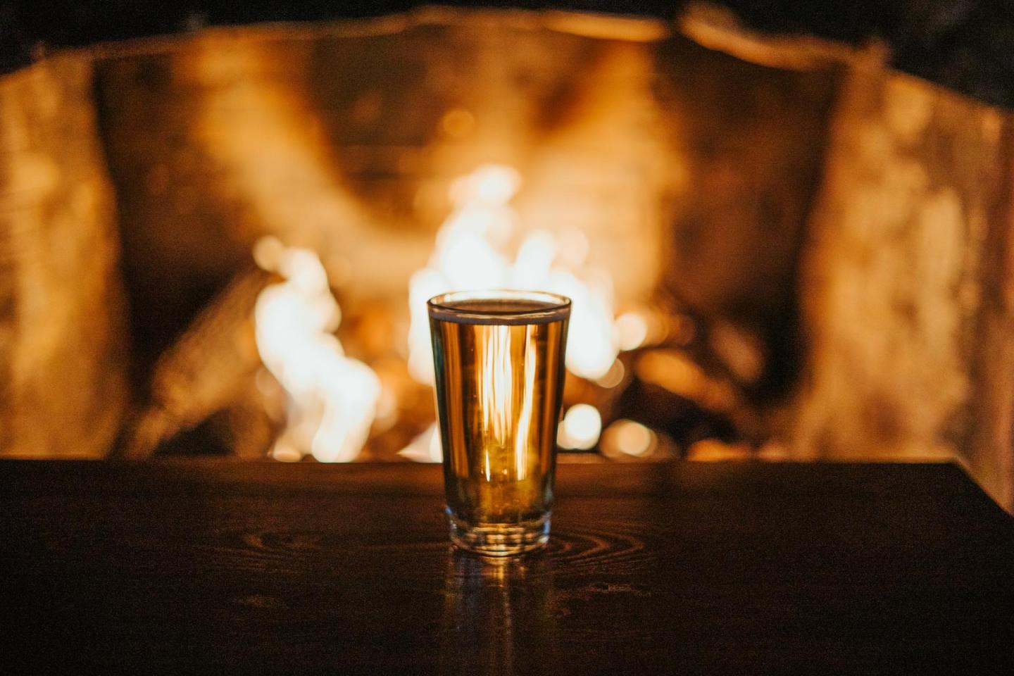 A glass of amber liquid on a table in front of a cozy fireplace.