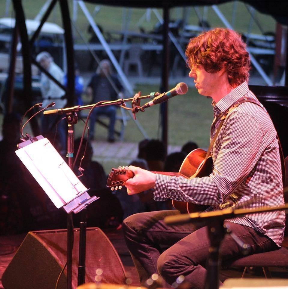 Musician playing guitar and singing on stage, seated under red lighting.