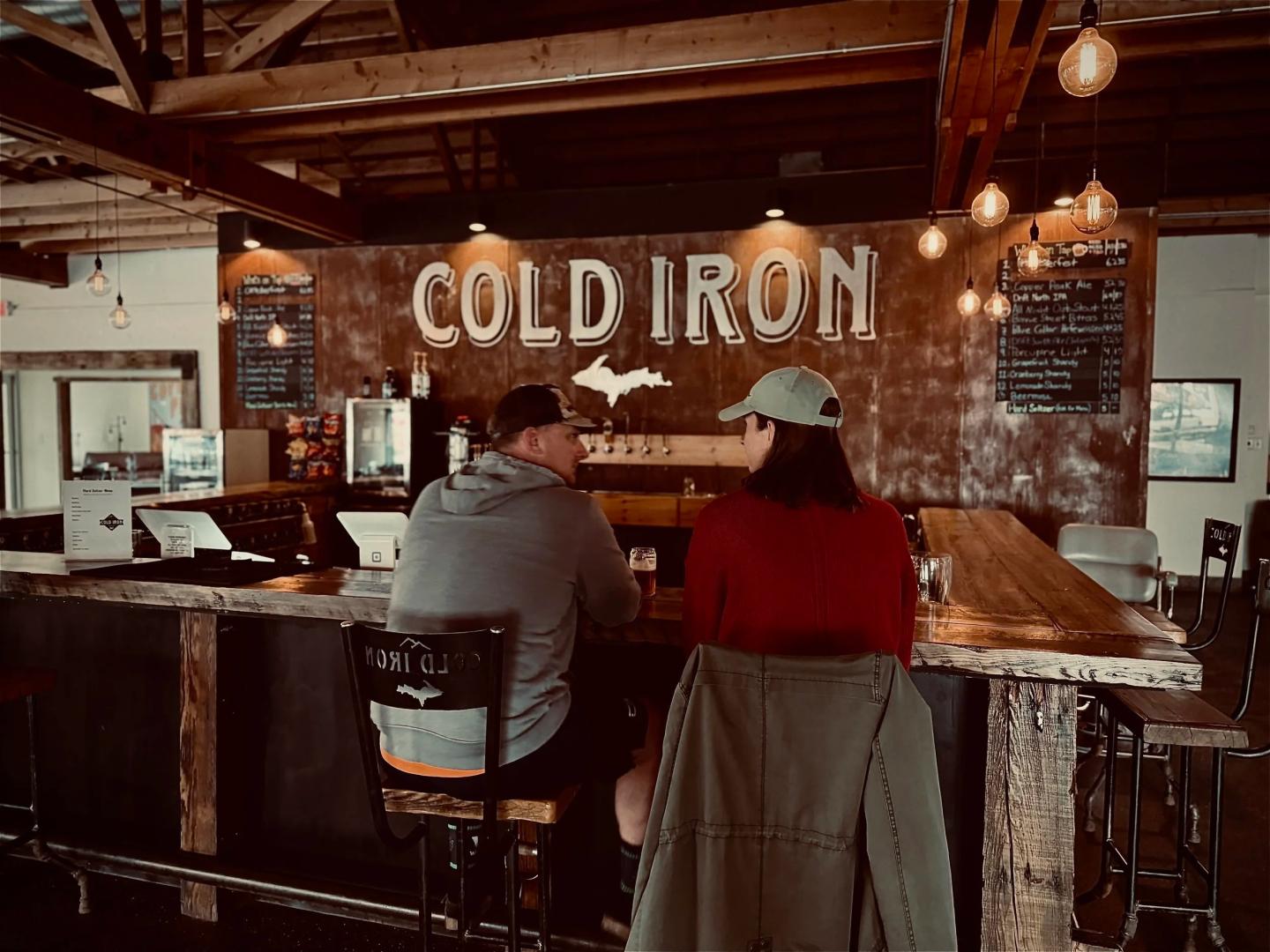 Two people seated at a rustic bar with "Cold Iron" sign above.