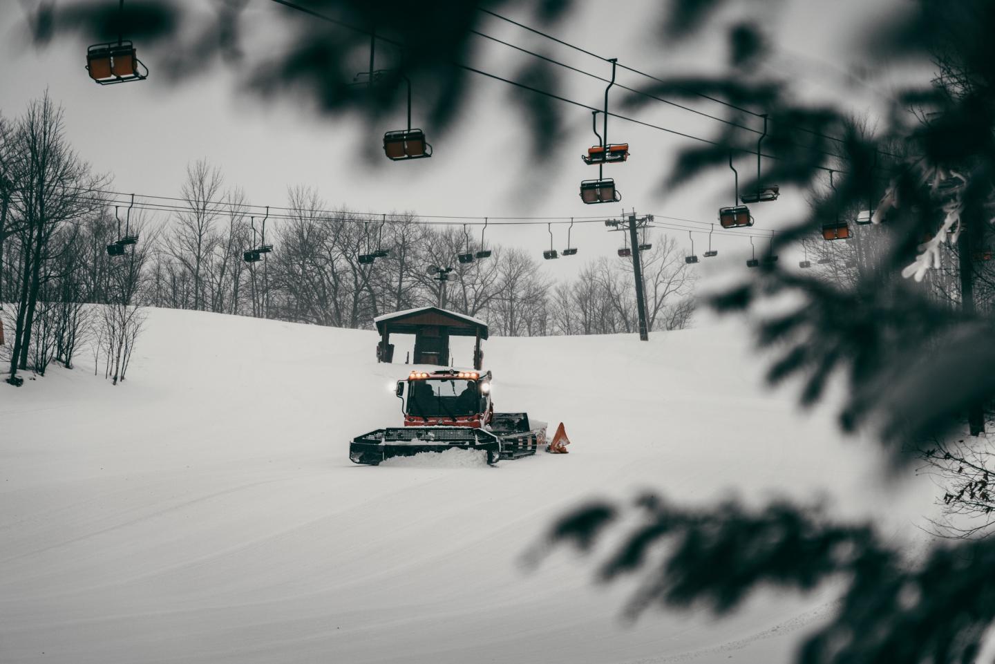 Snow groomer on a snowy slope, surrounded by bare trees, with a ski lift overhead.
