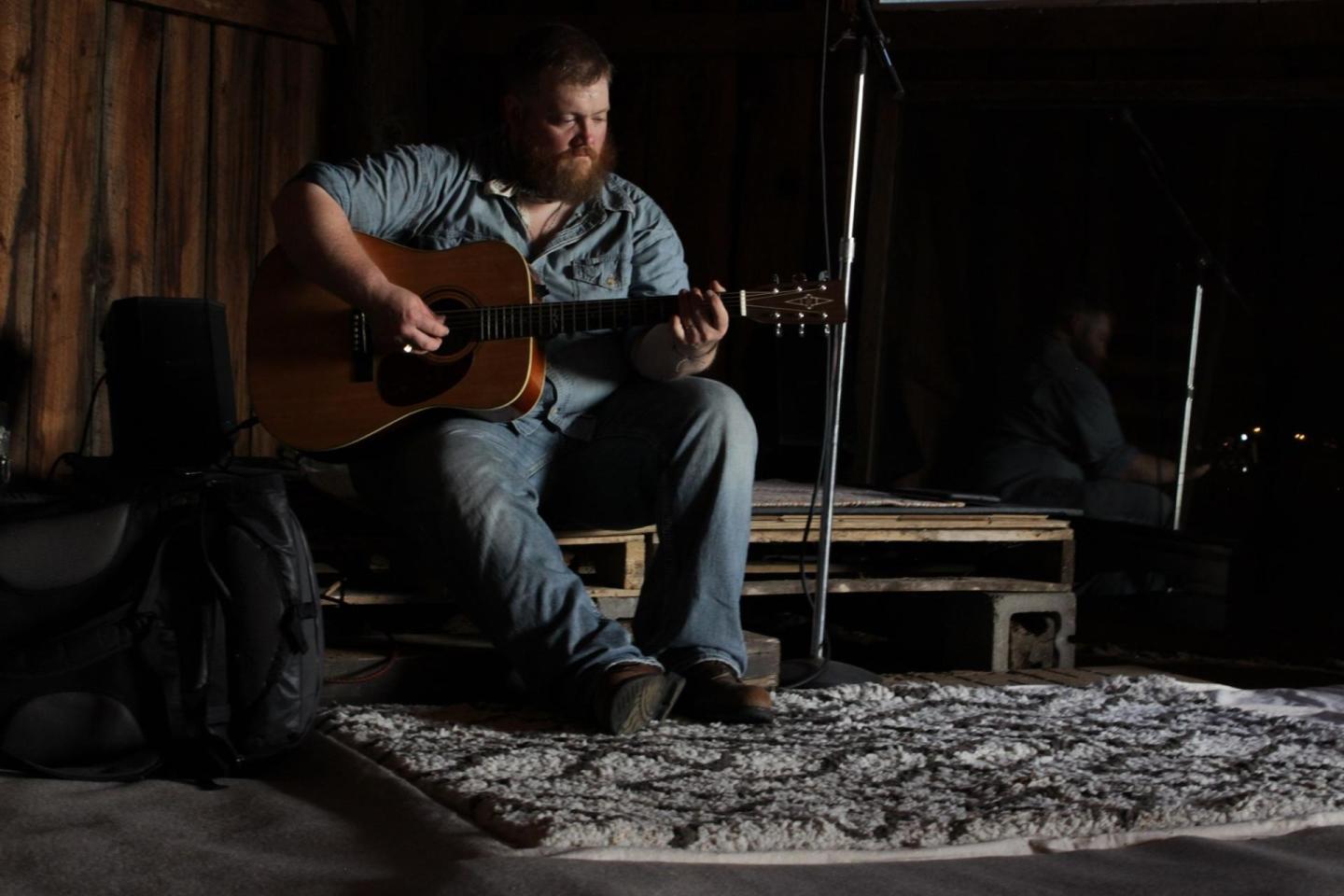 Man sitting on a stage, playing an acoustic guitar in dim light.
