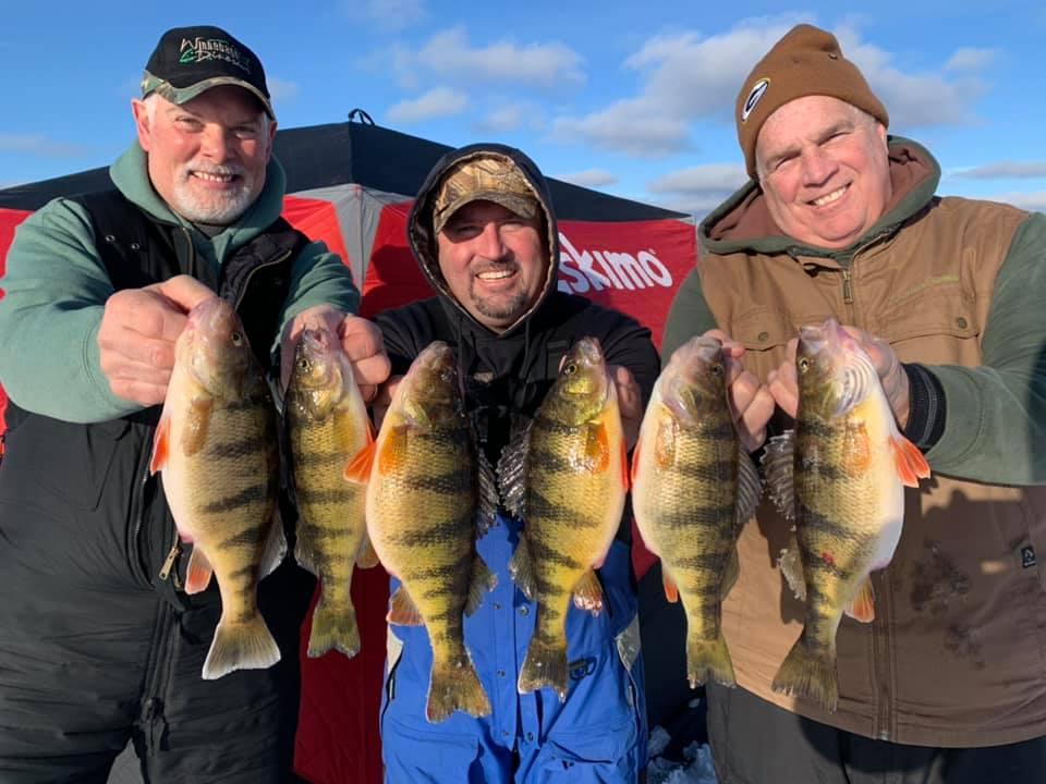 Three men holding large fish, smiling in front of a red ice fishing shelter.
