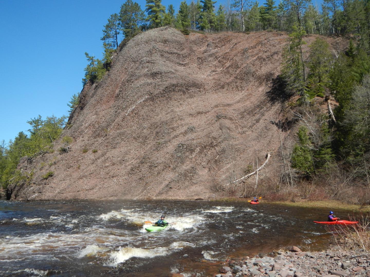 Kayakers paddle near a rocky cliff with trees on top under a clear blue sky.