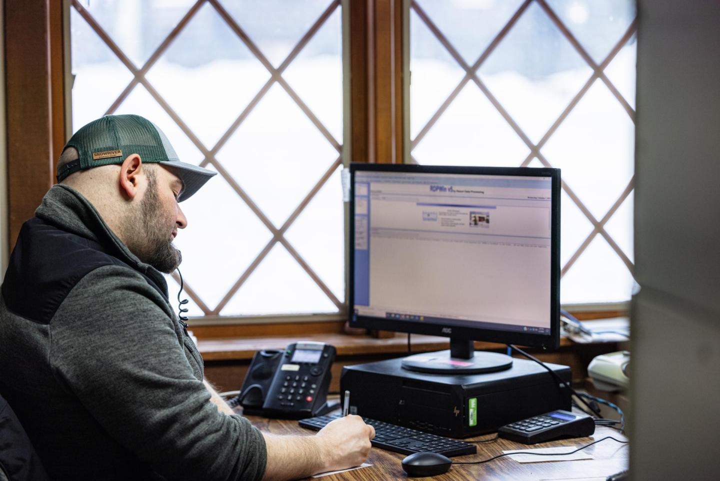 Man in baseball cap working at a desk with a computer and phone.
