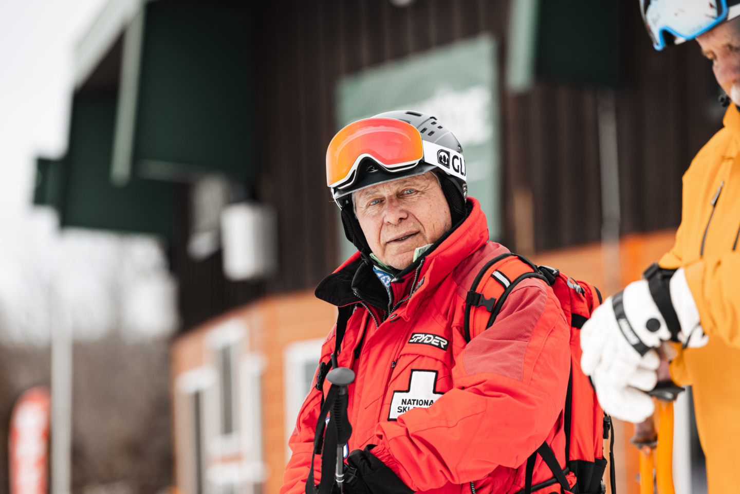 Ski patroller in red jacket with backpack, standing near a building.