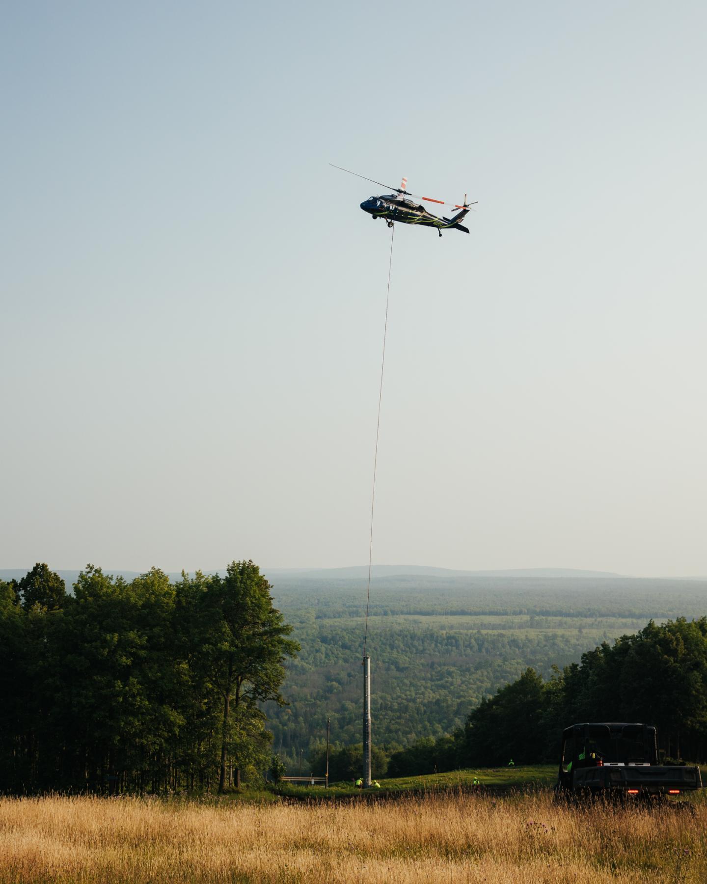 Helicopter in the sky over a field, lowering cable near forest edge.