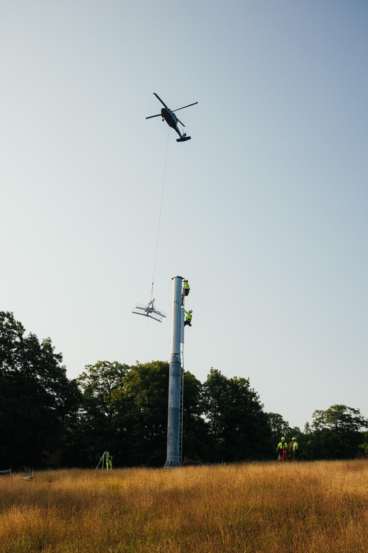 Helicopter lifting a structure above a field with trees in the background.