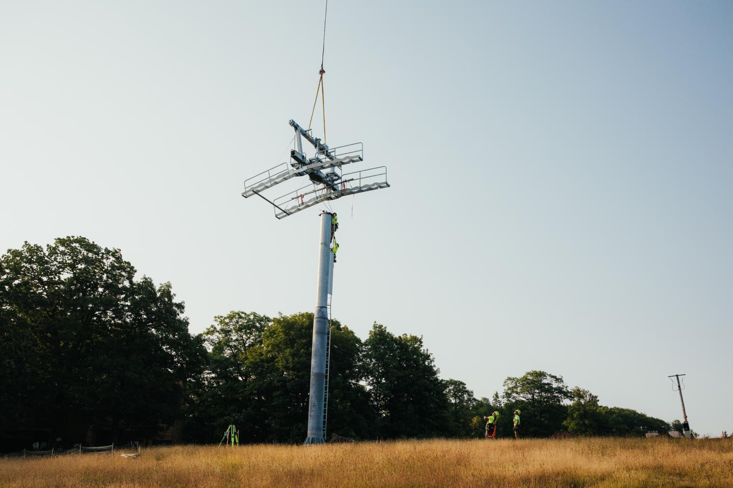 Chairlift tower installation in a grassy field with trees in the background.