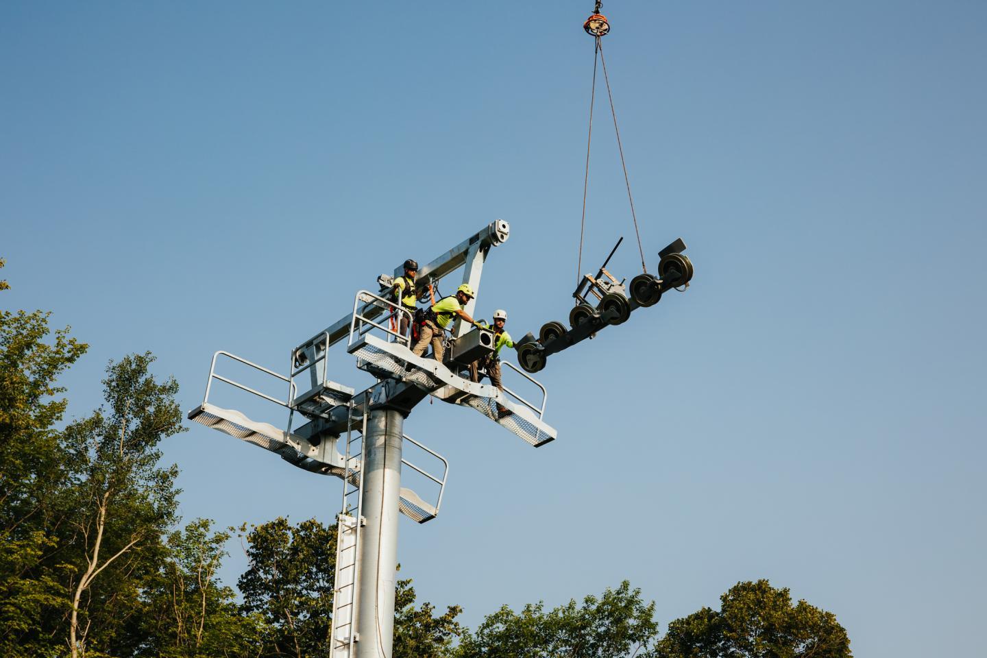 Workers on a ski lift tower with a crane overhead, clear sky.