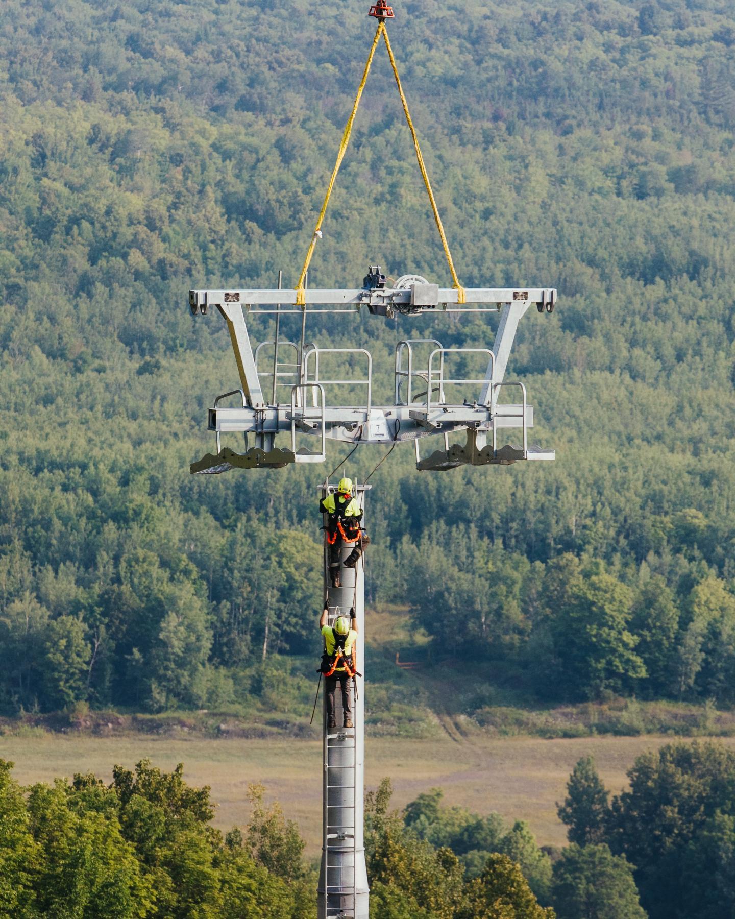 Workers installing a ski lift tower in a green, forested area.