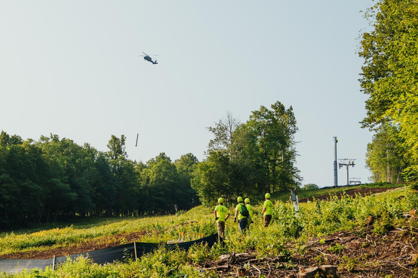 A helicopter flies over a green hillside with people below.