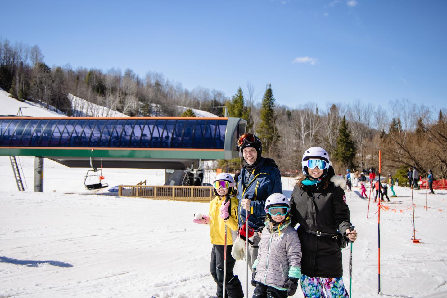 Family in winter gear stands on a snowy ski slope with trees and lift in the background.