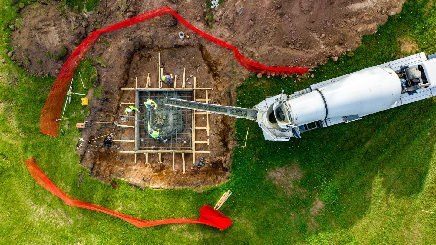 Concrete truck pouring into a wooden foundation frame surrounded by red fencing.