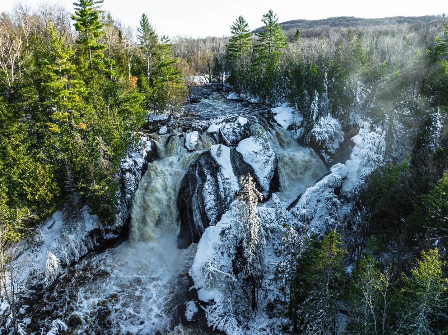Waterfall cascading between snowy rocks and surrounded by trees.