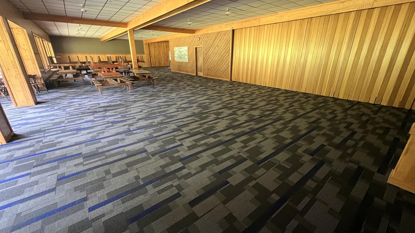 Large room with patterned carpet and wooden walls, empty chairs and tables on the left.
