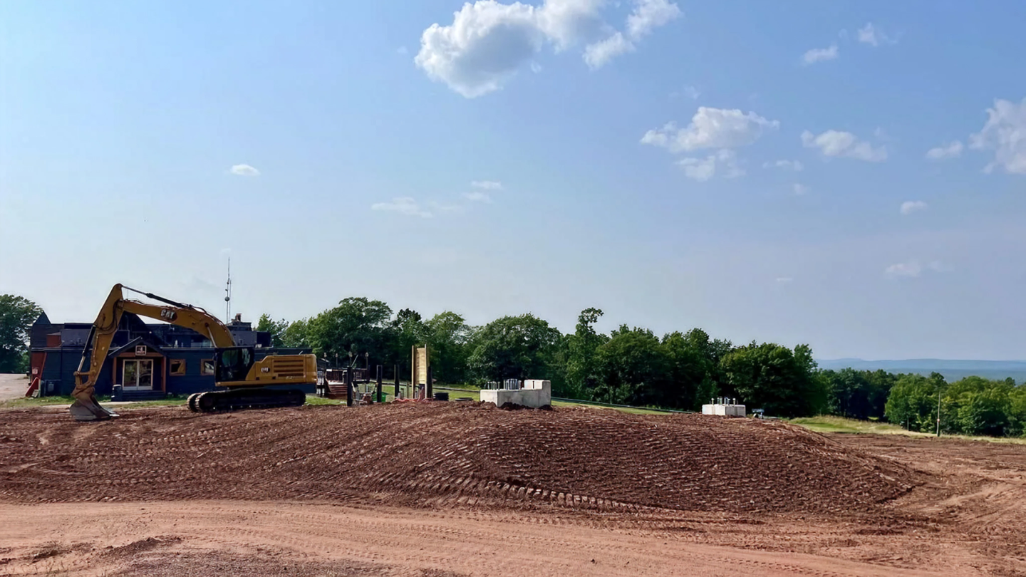 Construction site with an excavator on a dirt hill, surrounded by trees under a blue sky.