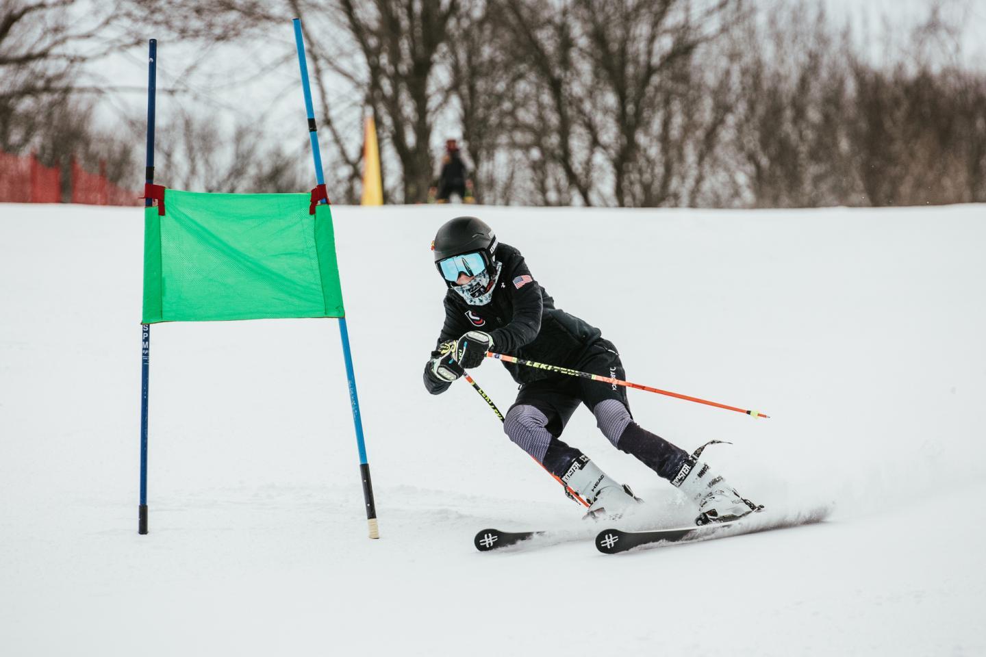 Skier racing downhill past a green gate, snow and trees in the background.
