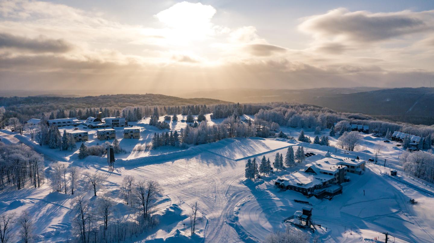 Snowy landscape with sun rays through clouds, buildings and trees blanketed in snow.
