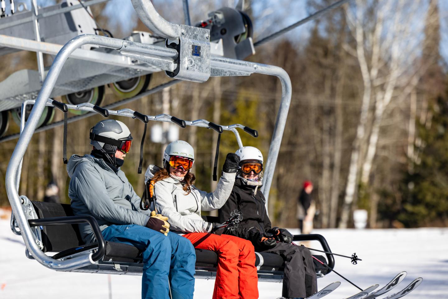 Skiers on a lift, wearing helmets and goggles, with snowy trees in the background.