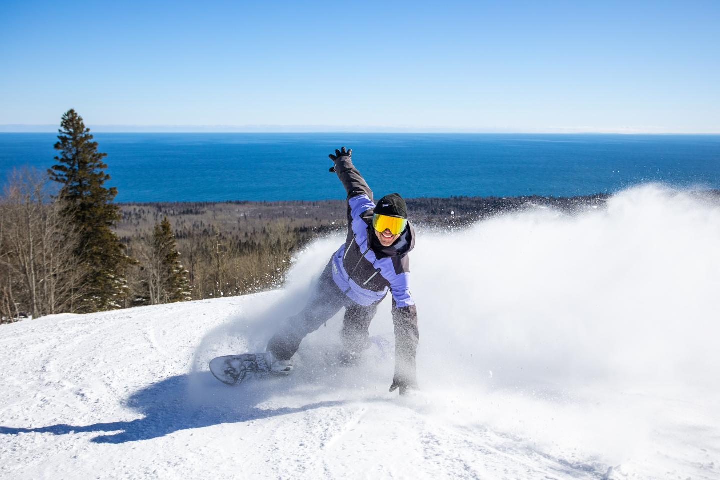 Snowboarder carving down a sunny slope with a blue ocean backdrop.