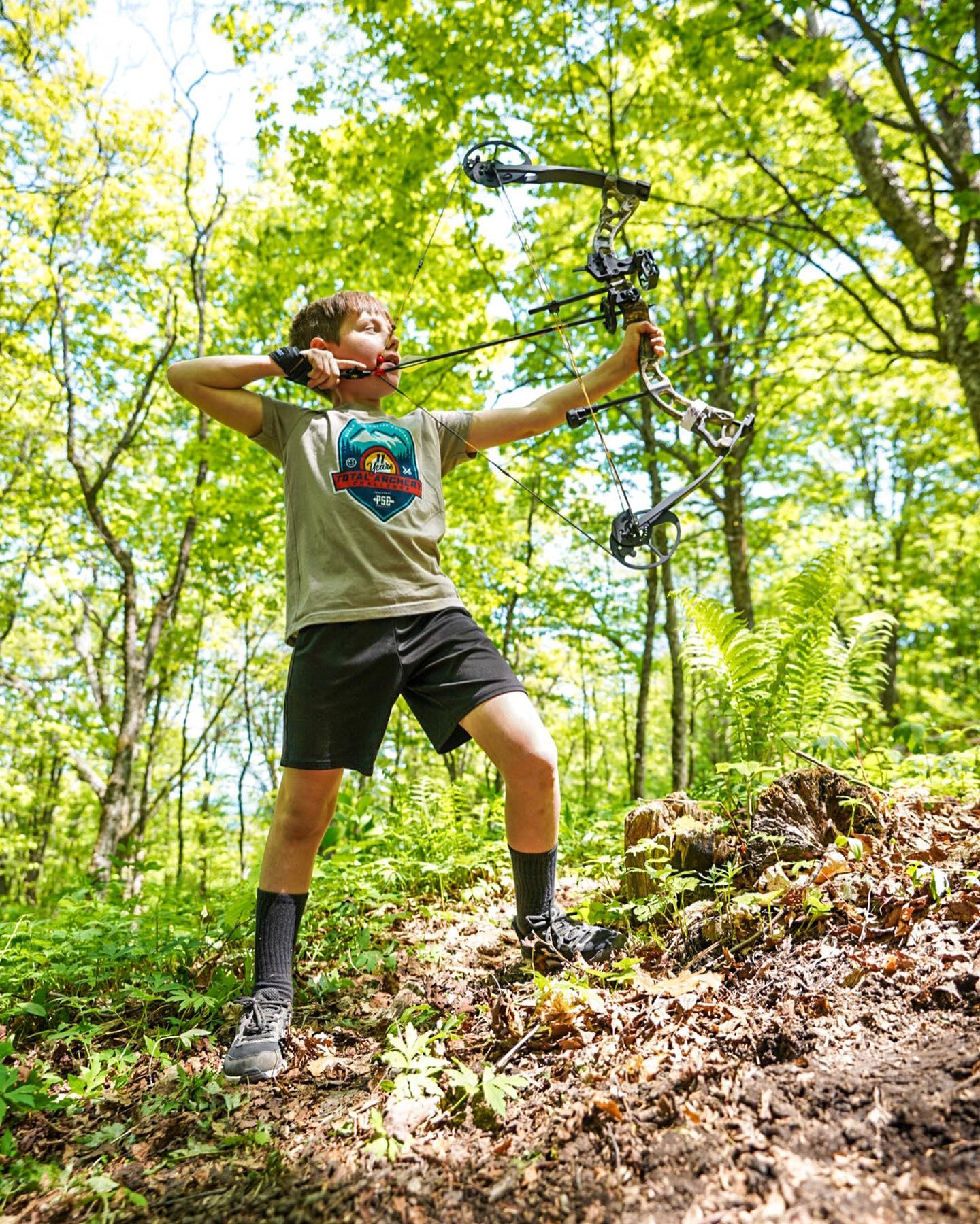 Boy aiming a bow and arrow in a sunlit forest.