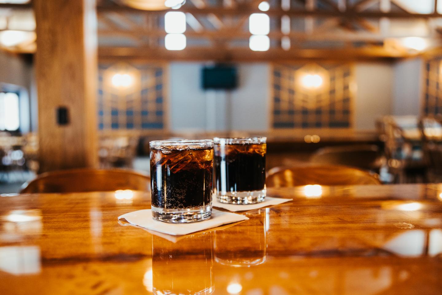 Two glasses of dark soda on a wooden bar, warm lighting in background.