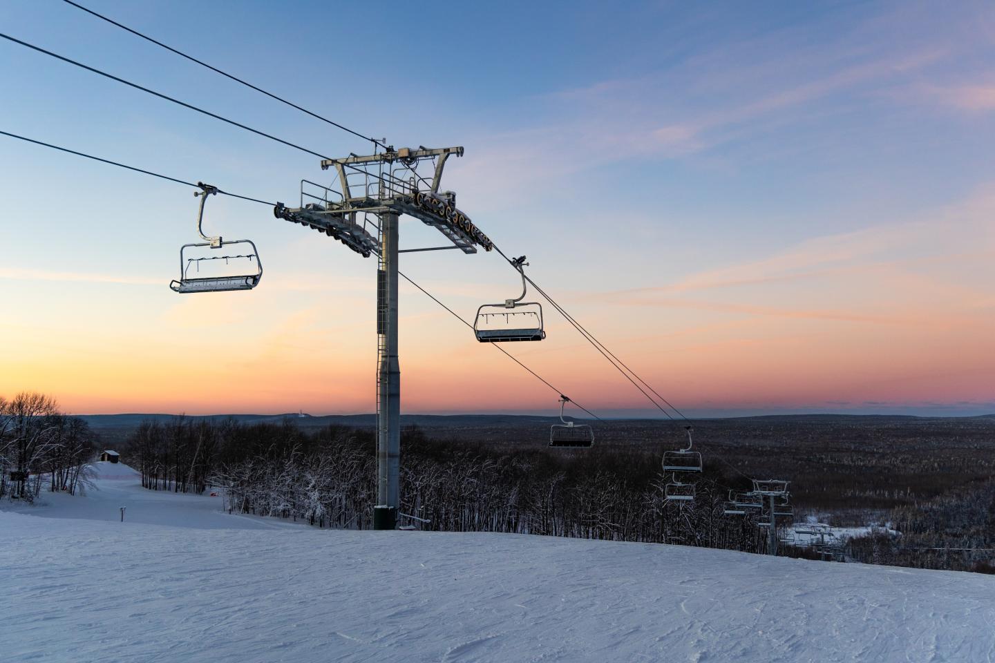 Ski lift over snowy slope at sunset, with colorful sky and distant trees.