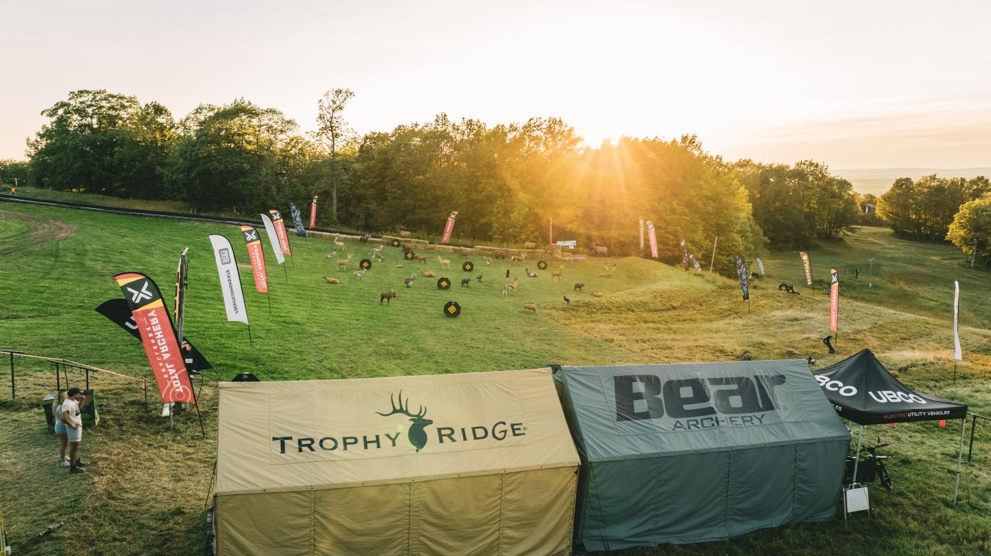 Sunlit outdoor archery range with tents and flags amid green fields and trees.