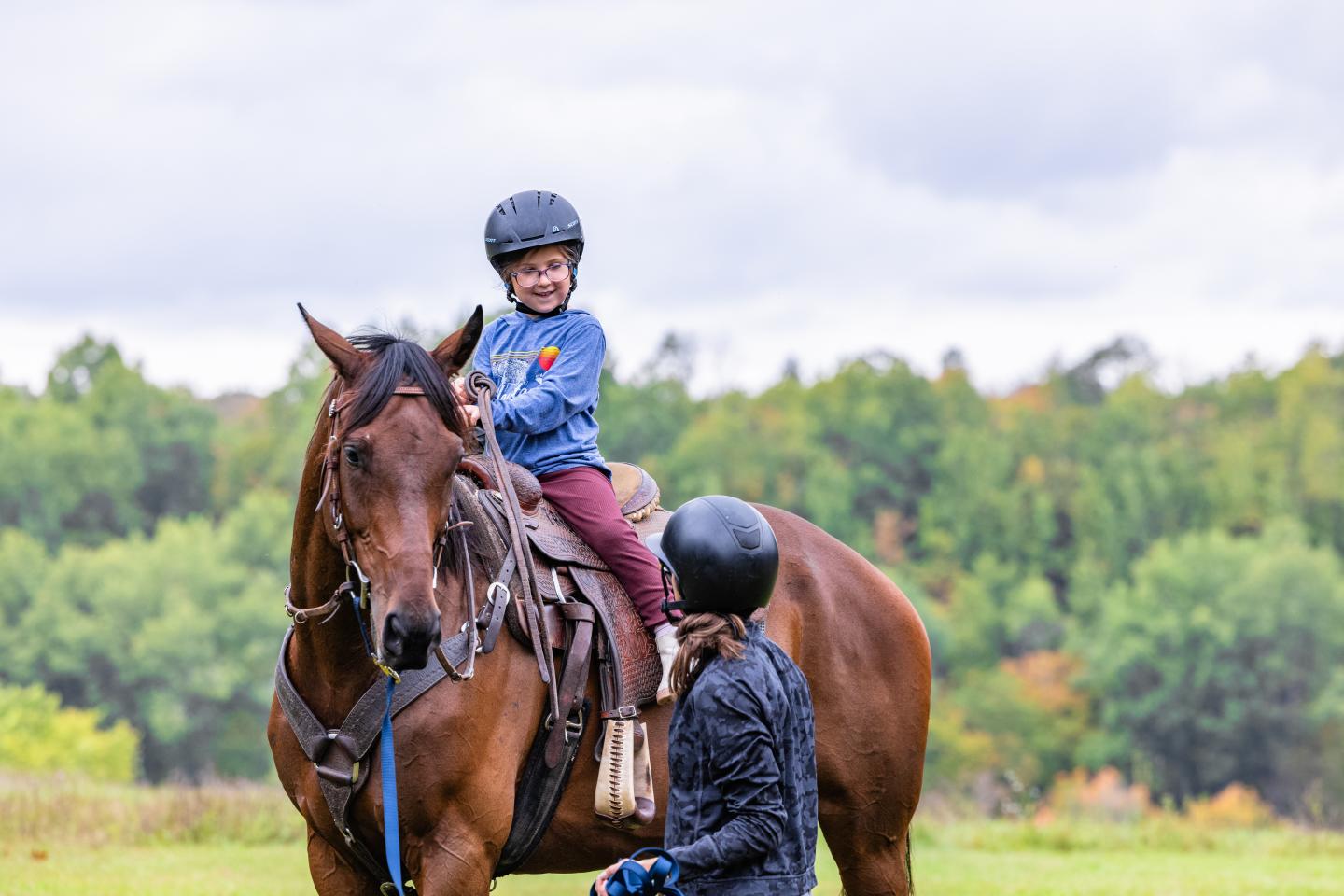 Child on horseback, smiling, with an adult beside. Lush green landscape in the background.