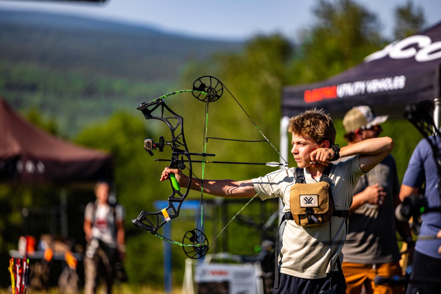 Archer aiming a compound bow outdoors, with tents and trees in the background.