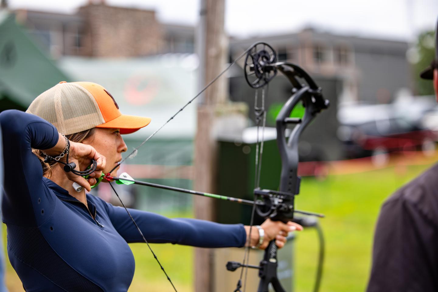 Archer in a cap aiming a compound bow outdoors.