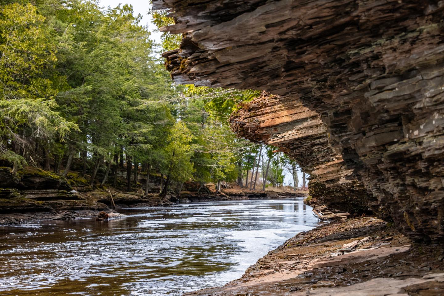 Rocky riverbank with overhanging cliff, flowing water, and lush green trees.