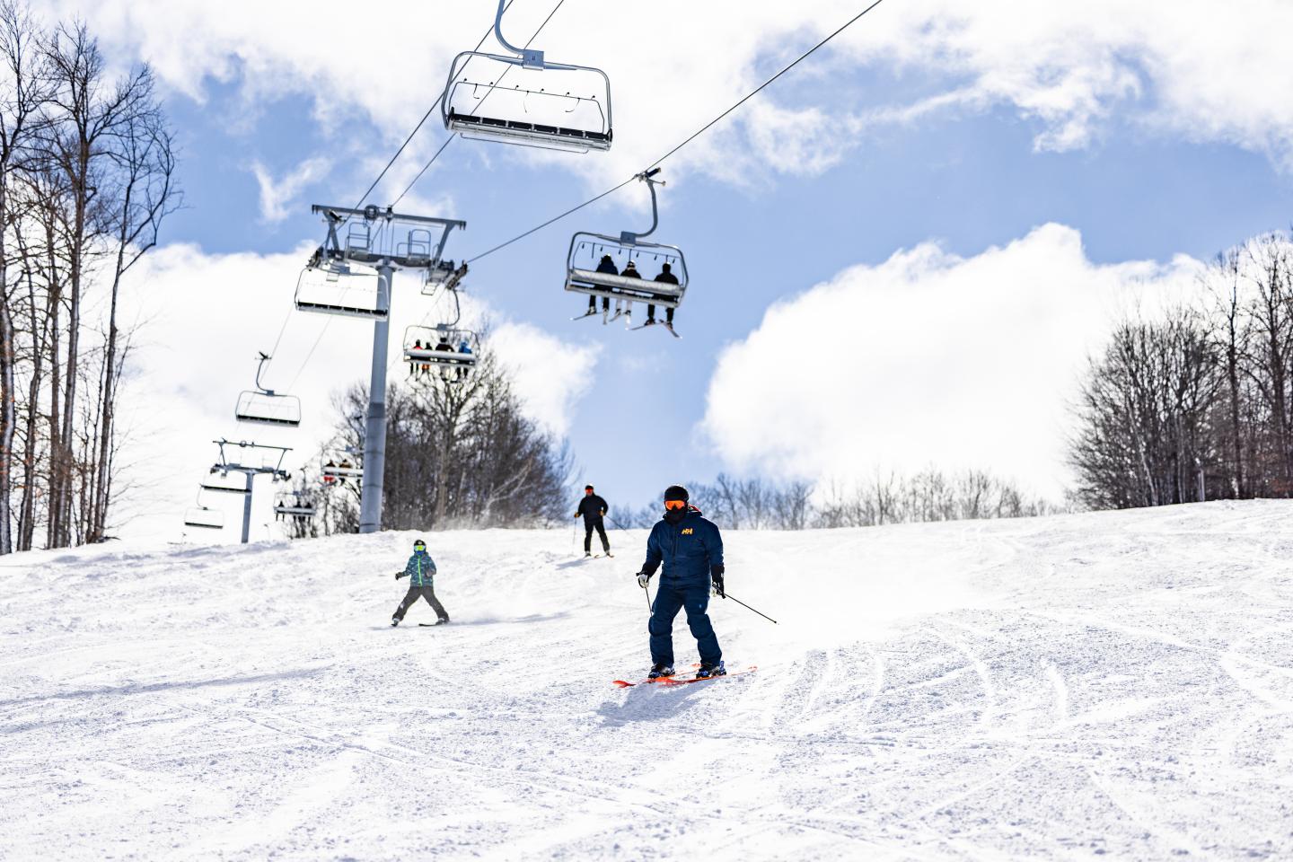 Skiers on a snowy slope with a ski lift above and clouds in the sky.
