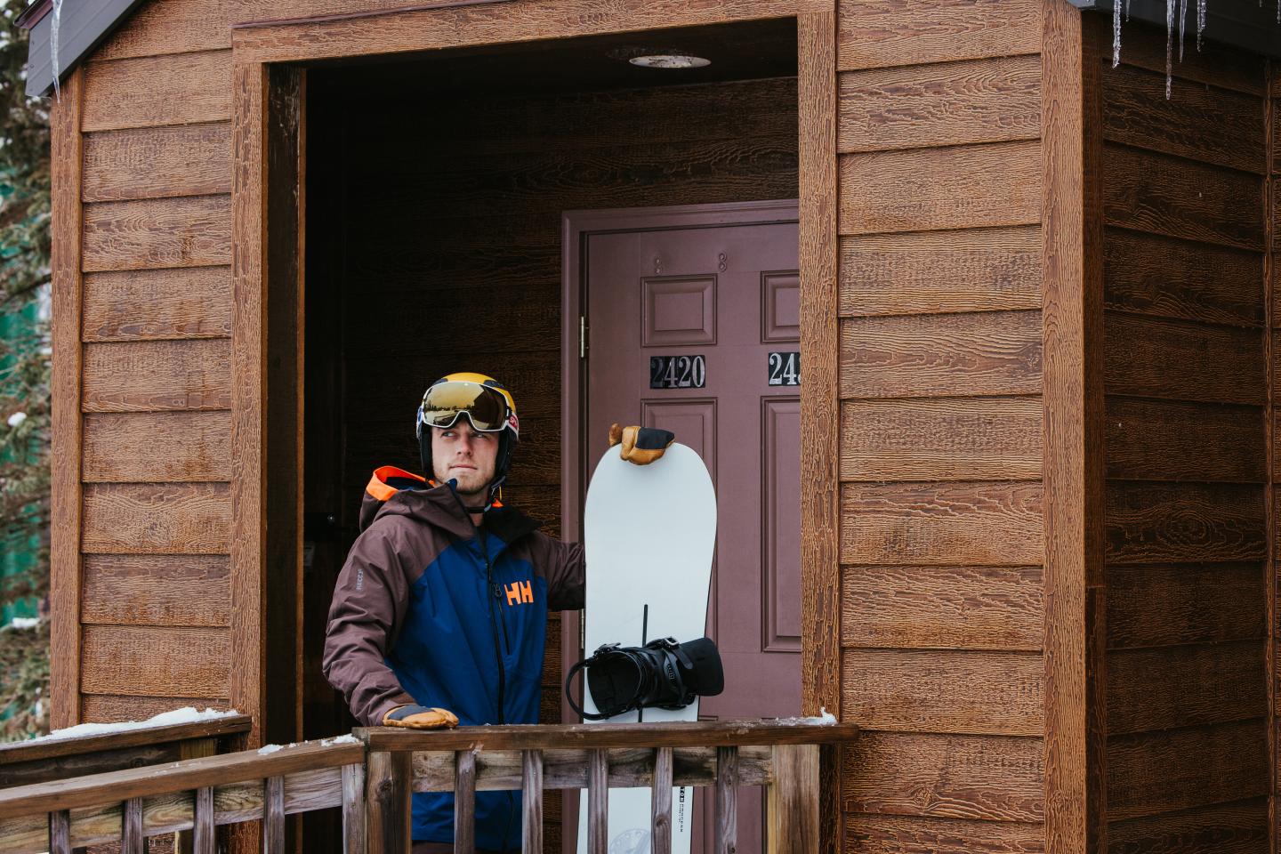 Snowboarder with gear stands at wooden cabin door.