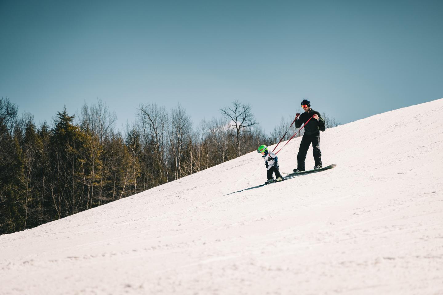 Adult teaching child to ski on snowy slope, clear sky.
