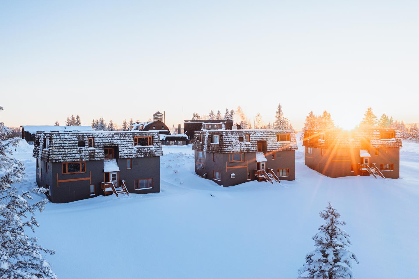 Snow-covered cabins at sunrise, surrounded by trees.