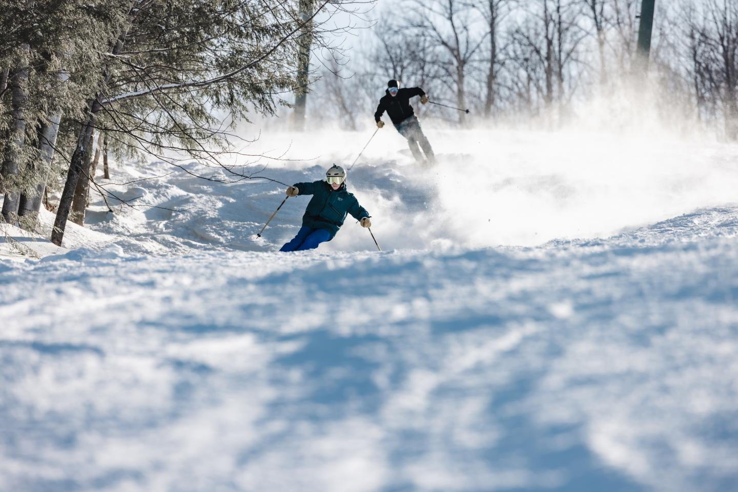 Two skiers on a snowy slope, trees in the background.