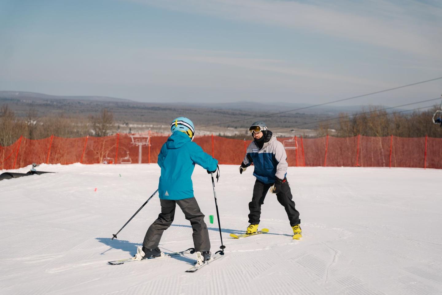 Two people skiing on a snowy slope, mountains in the background.