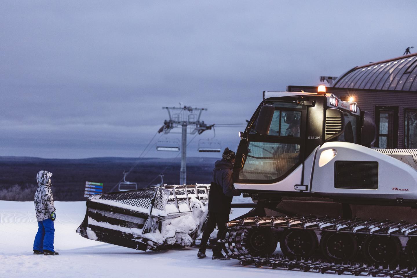 Snowcat grooming machine beside a person on a snowy slope at dusk.
