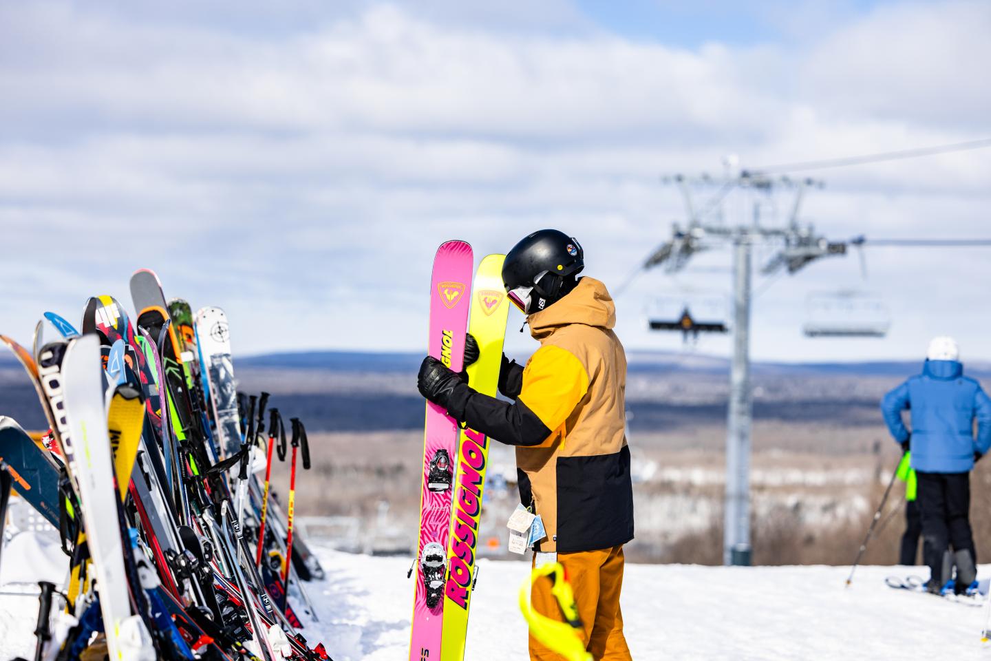 Skier in yellow pants holds pink skis beside a snowy trail.